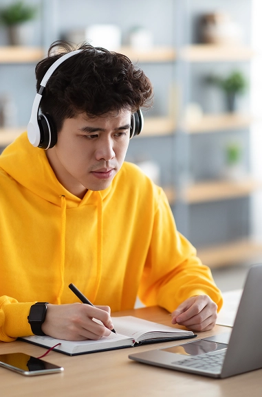 Young man wearing headphones and taking notes while studying on a laptop at a desk.